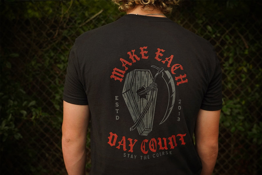 Person wearing a black t-shirt with red and gray text and graphic design on the back, standing against a chain-link fence.