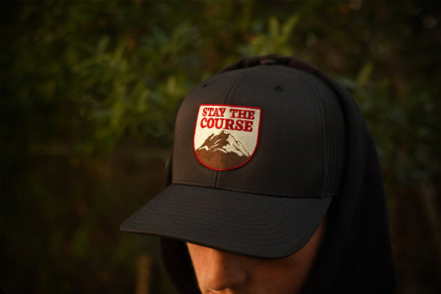 A person wearing a hat with a shield-shaped embroidered patch that reads 'STAY THE COURSE' above a mountain graphic, photographed outdoors with blurred green foliage in the background.