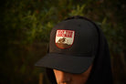 A person wearing a hat with a shield-shaped embroidered patch that reads 'STAY THE COURSE' above a mountain graphic, photographed outdoors with blurred green foliage in the background.