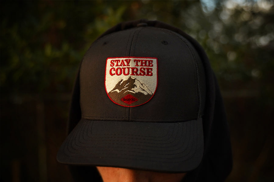 Person wearing a black trucker hat with patch style embroidery of a mountain with stay the course