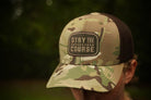Camouflage cap with 'Stay the Course' patch worn by a person outdoors