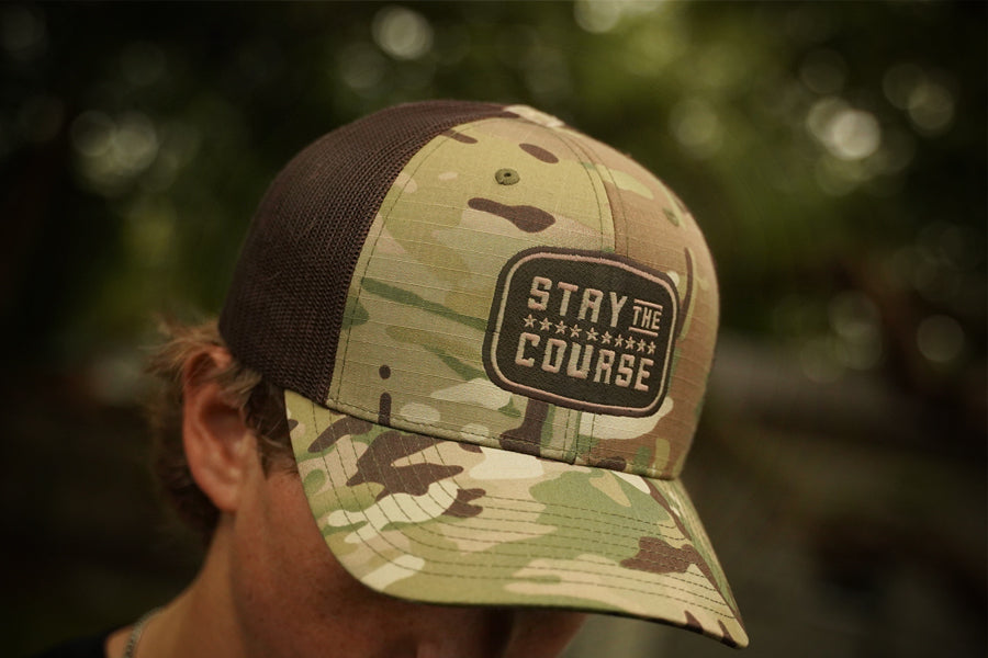 Camouflage cap with 'Stay the Course' logo worn by a person outdoors