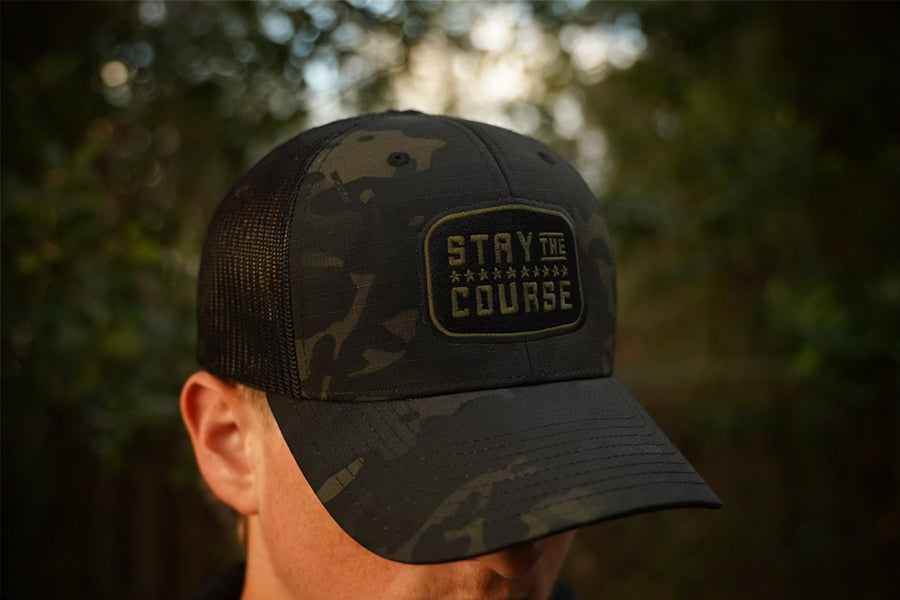 Person wearing a multicam black tactical trucker hat with "Stay the Course" embroidered in black and green, in an outdoor setting
