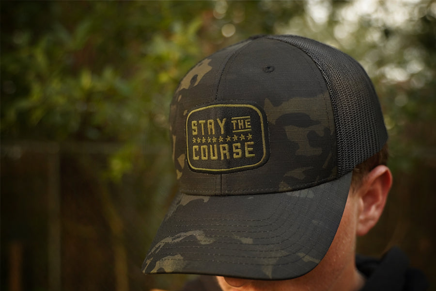 Person wearing a multicam black tactical trucker hat with "Stay the Course" embroidered in black and green, in an outdoor setting