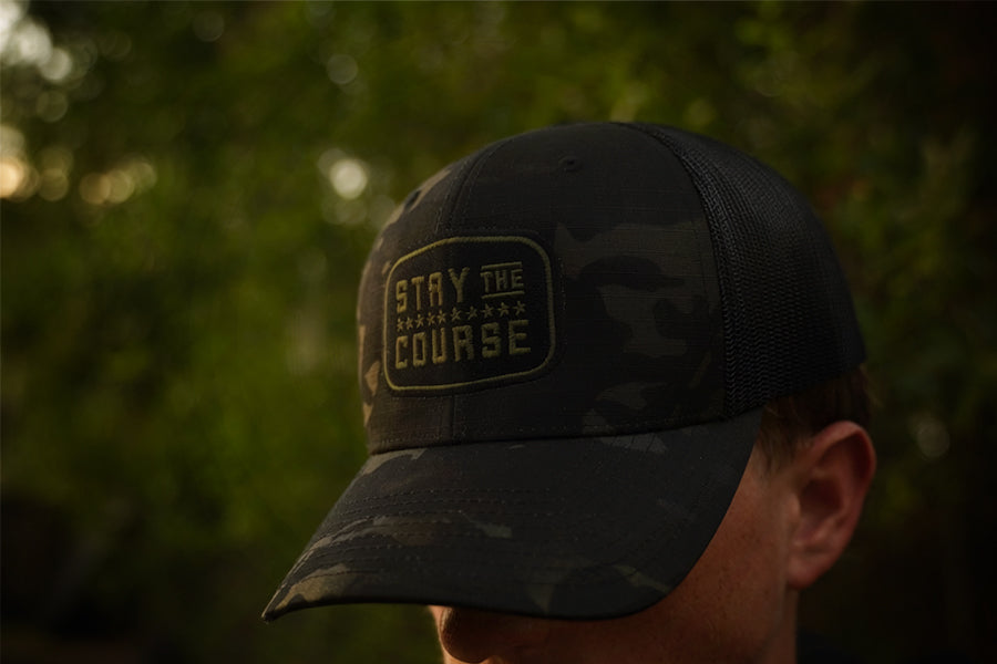 Person wearing a multicam black tactical trucker hat with "Stay the Course" embroidered in black and green, in an outdoor setting