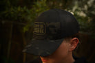 Person wearing a multicam black tactical trucker hat with "Stay the Course" embroidered in black and green, in an outdoor setting