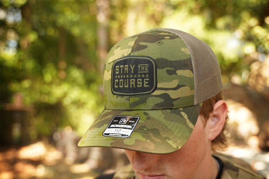 Camouflage cap with 'Stay the Course' patch worn by a person in a forest setting