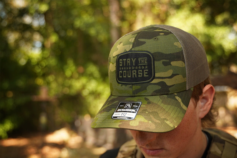 Camouflage cap with 'Stay the Course' patch worn by a person in a forest setting