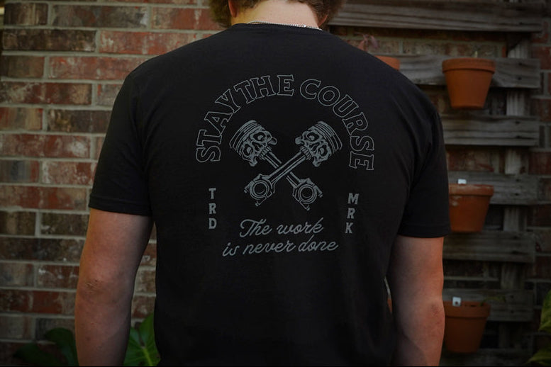 Person wearing a black t-shirt with a graphic design and text, standing in front of a brick wall.