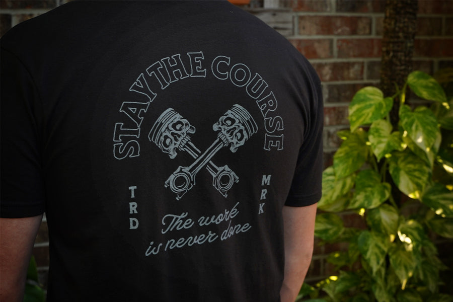 Black t-shirt with 'Stay the Course' graphic and text on the back, worn by a person standing against a brick wall.
