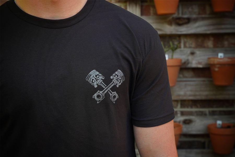 Person wearing a black t-shirt with a gray piston design, standing against a rustic wooden wall with potted plants.