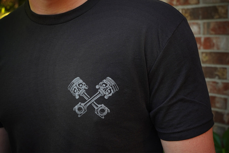 Black t-shirt with gray piston design, against a brick wall background.