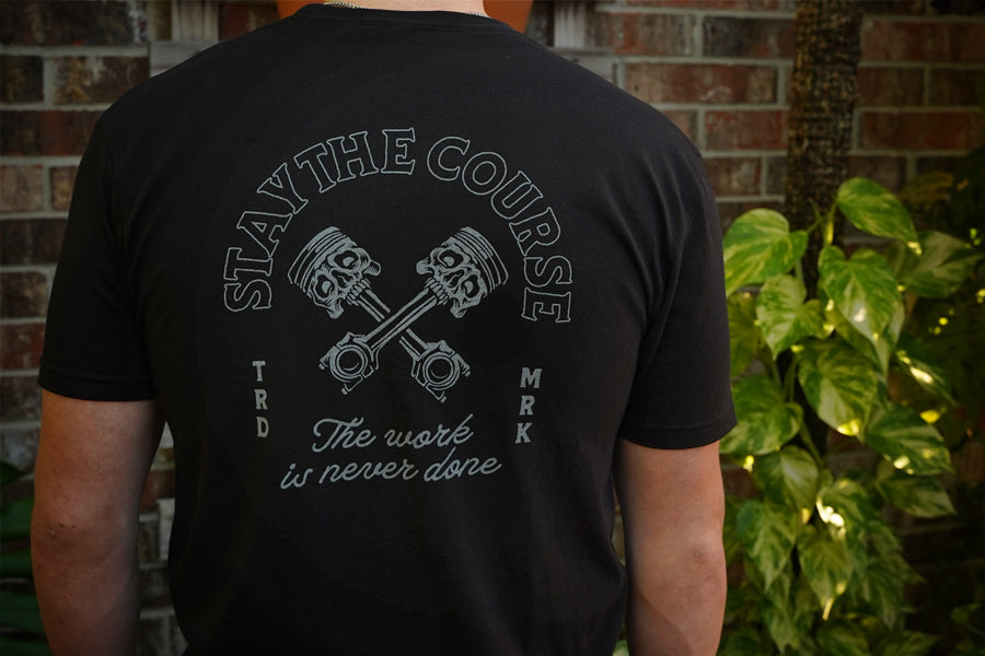 Person wearing a black t-shirt with a graphic design and text against a brick wall and plant background