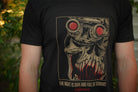 Person wearing a black t-shirt with tan and red graphic print and the words "the night is dark and full of terrors", in front of greenery