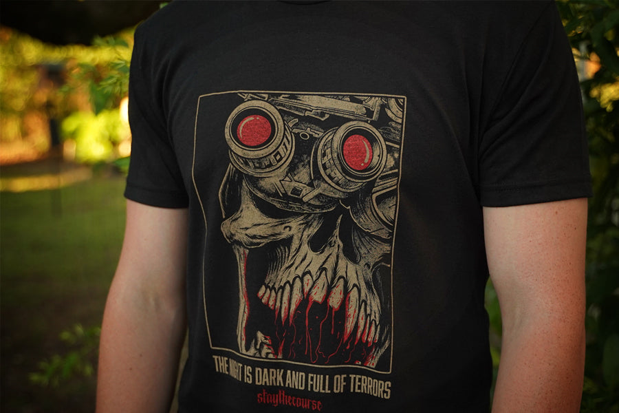 Person wearing a black t-shirt with tan and red graphic print and the words "the night is dark and full of terrors", in an outdoor setting.