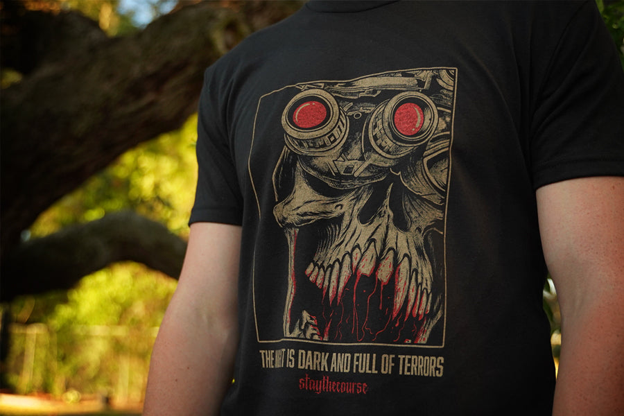 Person wearing a black t-shirt with tan and red graphic print and the words "the night is dark and full of terrors", standing in front of a tree