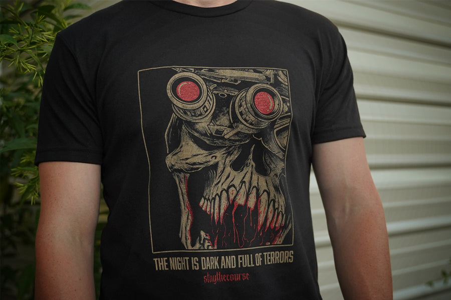 Person wearing a black t-shirt with tan and red graphic print and the words "the night is dark and full of terrors", standing in front of a building.