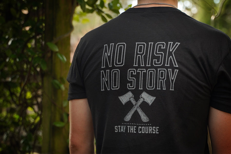 Person wearing a black t-shirt with 'No Risk No Story, Stay the Course' text and graphic on the back, standing in a forest.