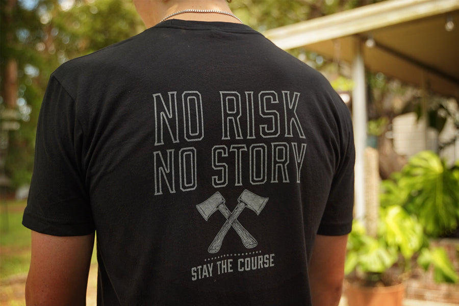 Person wearing a black t-shirt with 'No Risk No Story Stay the Course' text and graphic outdoors.