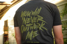 Person wearing a black t-shirt with green graphic print that says "you will never outwork me, stay the course", standing in front of a building.