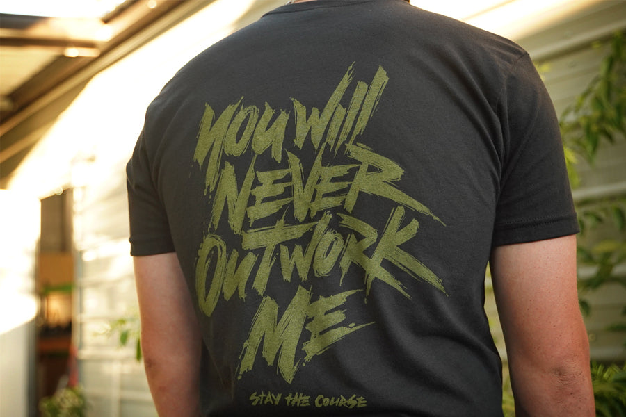 Person wearing a black t-shirt with green graphic print that says "you will never outwork me, stay the course", standing in front of a building.