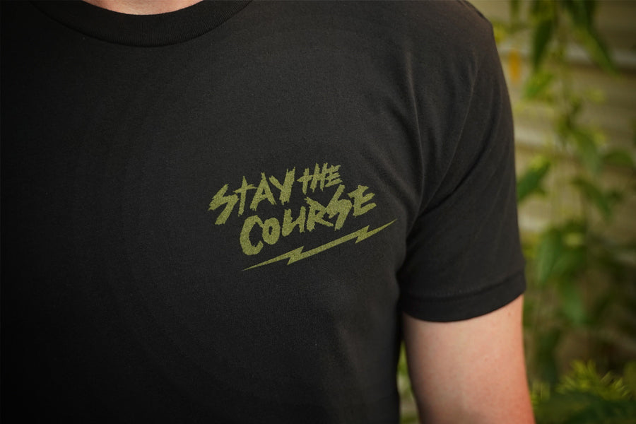 Person wearing a black t-shirt with green graphic print that says "stay the course", standing in front of a building.