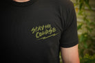 Person wearing a black t-shirt with green graphic print that says "stay the course", standing in front of a building.
