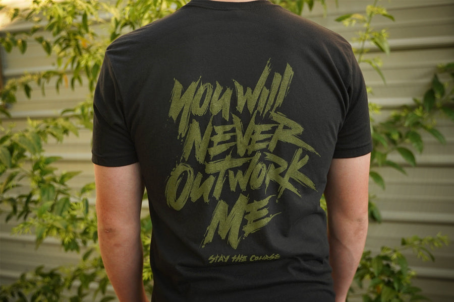 Person wearing a black t-shirt with green graphic print that says "you will never outwork me, stay the course", standing in front of a building.