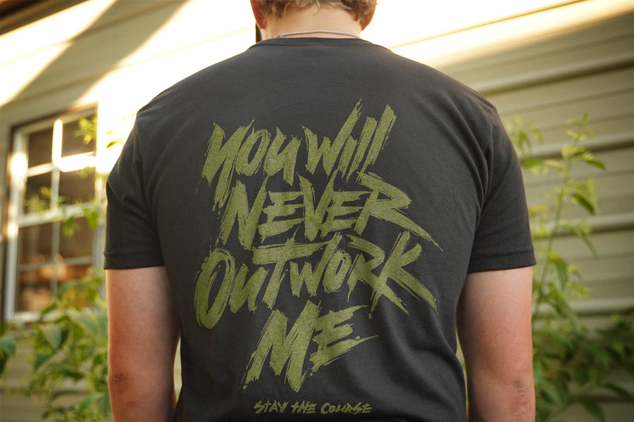 Person wearing a black t-shirt with green graphic print that says "you will never outwork me, stay the course", standing in front of a building.