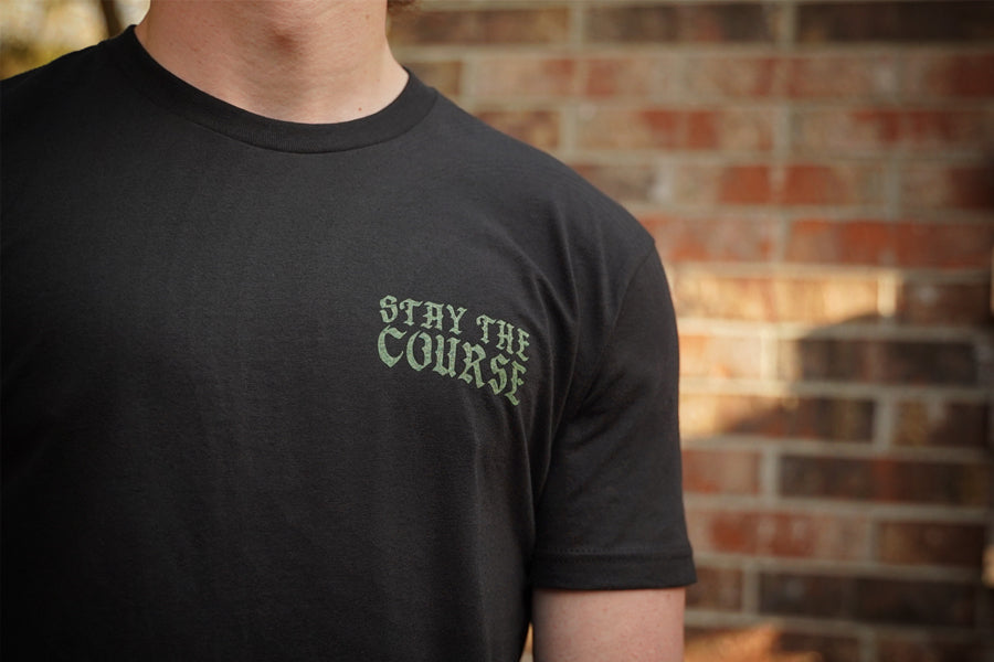 Person wearing a black t-shirt with 'Stay the Course' text against a brick wall.