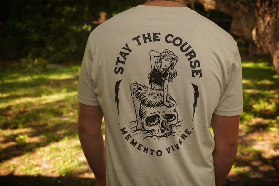 Person wearing a beige t-shirt with a graphic design and text 'Stay the Course Memento Vivere' outdoors.