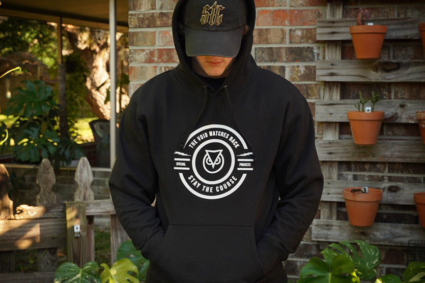 Person wearing a black hoodie with a logo and text, standing in front of a brick wall with potted plants.