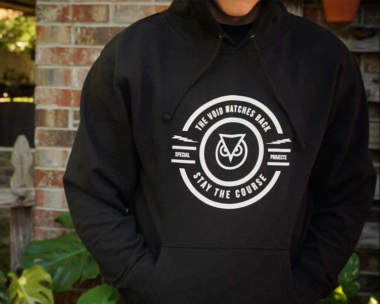 Person wearing a black hoodie with a white logo and text, standing in front of a brick wall.