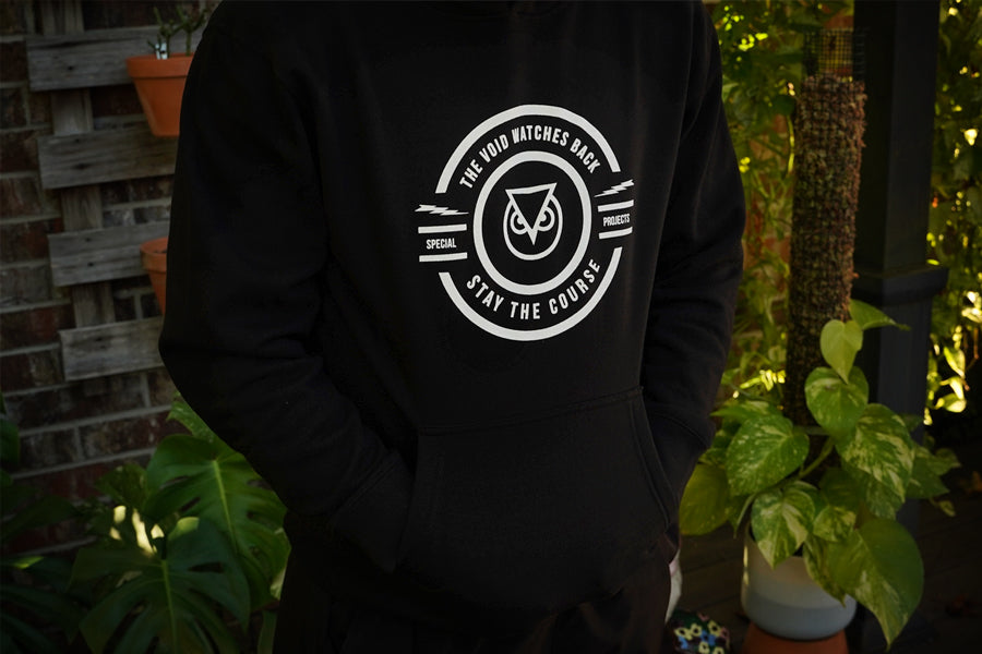 Black hoodie with a white logo and text worn by a person, set against a natural background with plants.