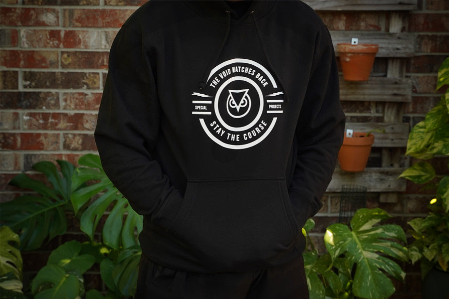 Person wearing a black hoodie with a white logo against a brick wall and plant background