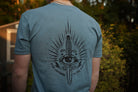 Person wearing a blue t-shirt with a black eye and sword design on the back, standing outdoors.