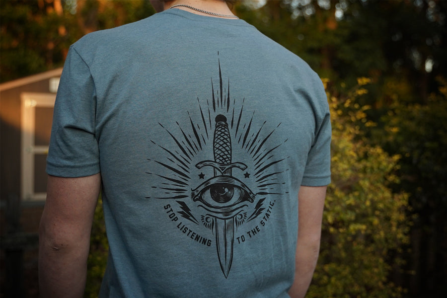 Person wearing a blue t-shirt with a black eye and sword design on the back, standing outdoors.