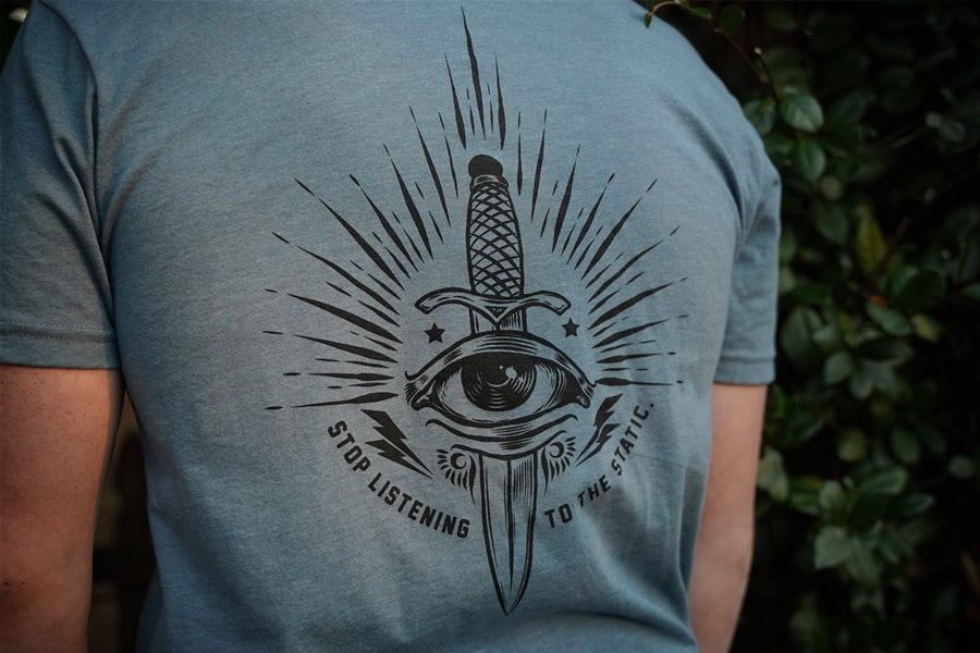 Person wearing a blue t-shirt with a black graphic design on the back, featuring an eye and a sword.