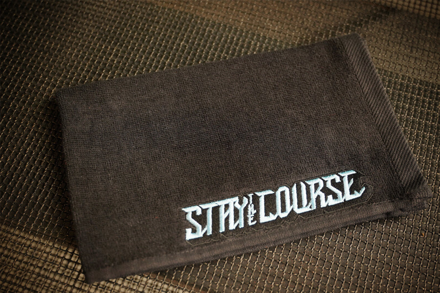 A black towel featuring the embroidered baby blue text 'STAY COURSE' in a stylized font, laid flat on a brown woven surface.