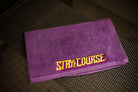 A purple towel with 'STAY ON COURSE' embroidered in yellow and purple resting on a dark textured surface.