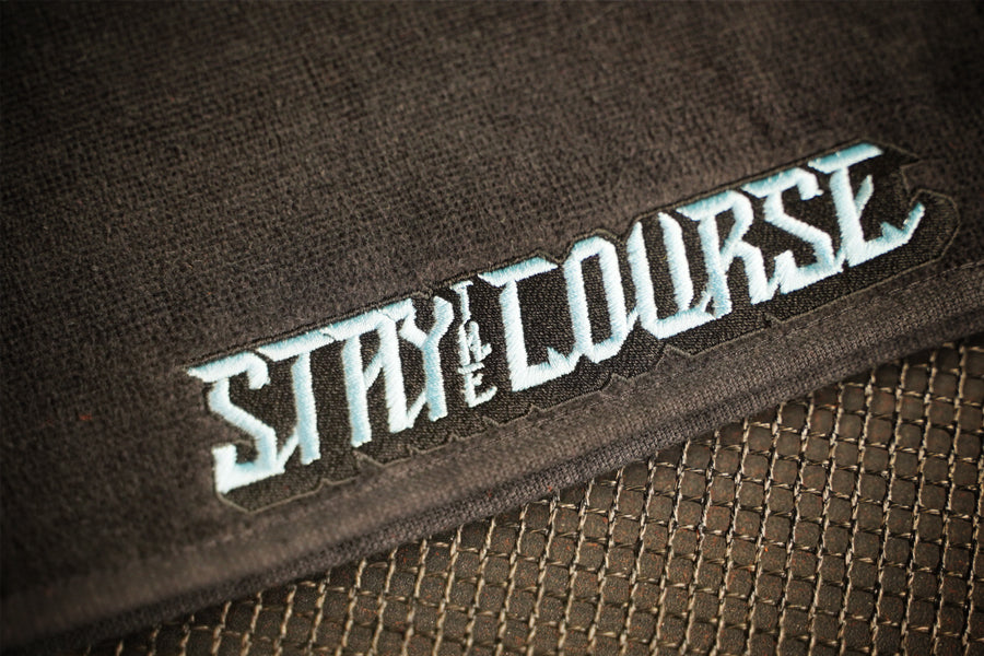 Close-up of embroidery with the words 'STAY ON THE COURSE' in stylized light blue lettering, placed over a dark mesh surface.