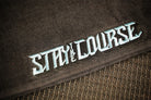 Close-up of embroidery with the words 'STAY ON THE COURSE' in stylized light blue lettering, placed over a dark mesh surface.