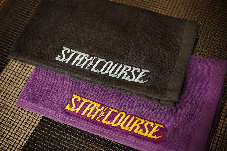 Two folded towels stacked on a woven mat: a black towel on top with baby blue embroidered 'STAY ON THE COURSE' text, and a purple towel underneath with yellow and purple embroidered 'STAY ON THE COURSE' text.