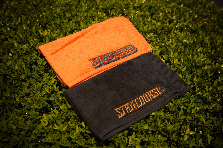 Two folded towels, one orange and one black, with 'Stay the Course' branding on a grassy background.