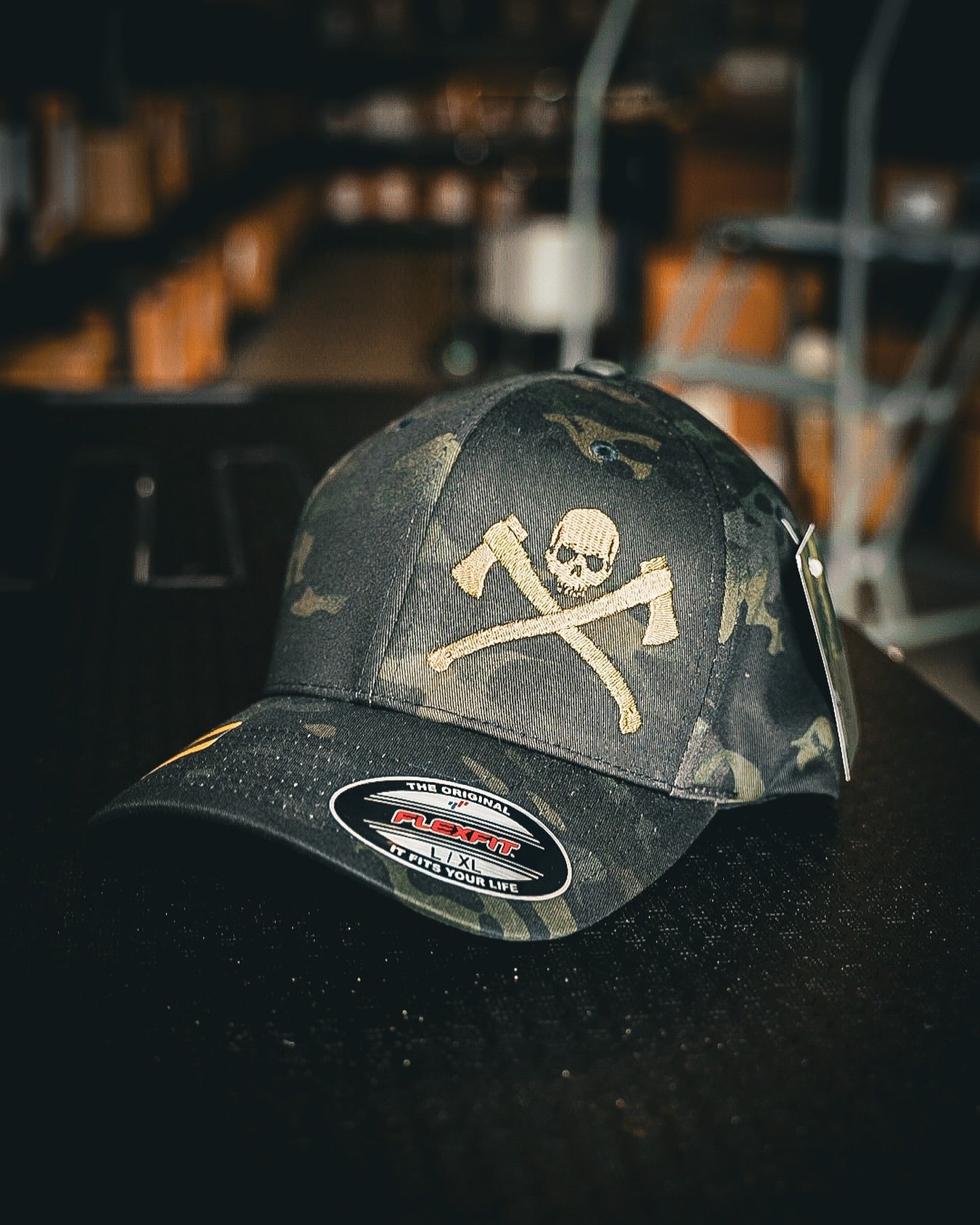 A dark camouflage baseball cap featuring a skull and crossed axes graphic on the front, resting on a dark surface with a blurred indoor background.