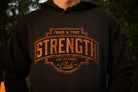 A close-up of a person wearing a black hoodie with a large rust colored graphic on the chest that reads 'TRIED & TRUE STRENGTH CLUB STAY THE COURSE'.