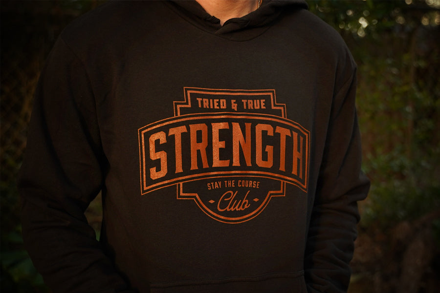 A person wearing a black hoodie with a large rust colored graphic on the chest that reads 'TRIED & TRUE STRENGTH CLUB' and 'STAY THE COURSE'. The background is a blurred outdoor scene with green foliage.
