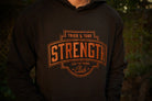 A person wearing a black hoodie with a large rust colored graphic on the chest that reads 'TRIED & TRUE STRENGTH CLUB' and 'STAY THE COURSE'. The background is a blurred outdoor scene with green foliage.