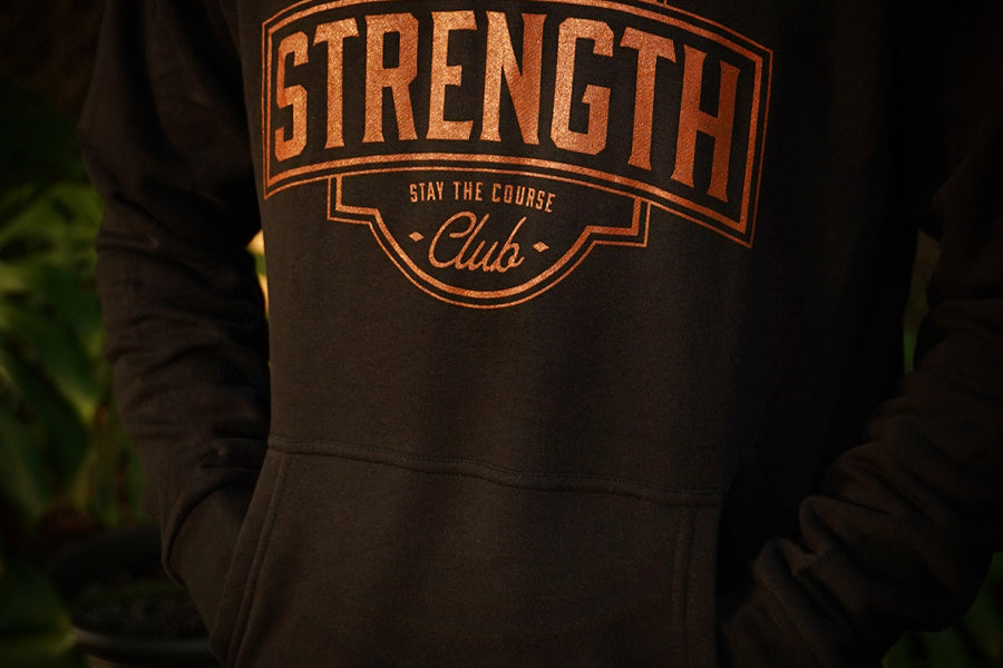 A close-up of a person wearing a black hoodie with a rust colored graphic print on the chest that reads 'STRENGTH Club' and 'STAY THE COURSE'.