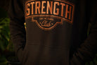 A close-up of a person wearing a black hoodie with a rust colored graphic print on the chest that reads 'STRENGTH Club' and 'STAY THE COURSE'.